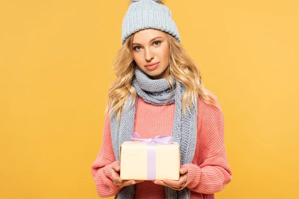 Atractiva mujer rubia en sombrero en bufanda sosteniendo caja de regalo aislada en amarillo - foto de stock