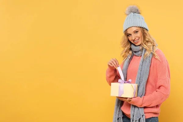 Smiling woman in hat and scarf holding ribbon of gift box isolated on yellow — Stock Photo
