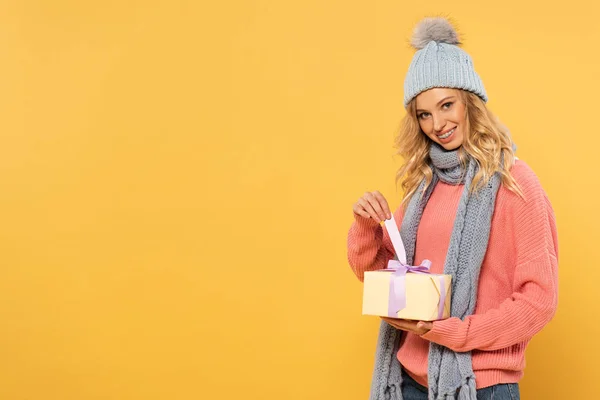 Mujer sonriente con sombrero y bufanda sosteniendo cinta de caja de regalo aislada en amarillo - foto de stock