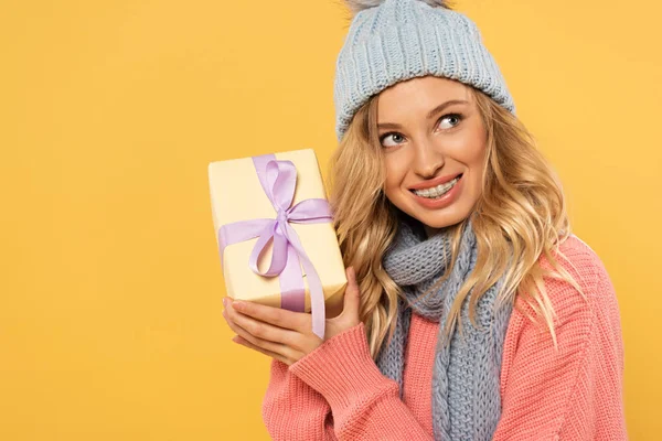 Lächelnde Frau mit Hut und Schal hält Geschenkbox isoliert auf gelb — Stockfoto