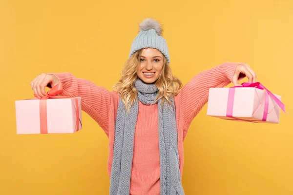 Femme blonde souriante en chapeau et écharpe tenant des boîtes-cadeaux isolées sur jaune — Photo de stock