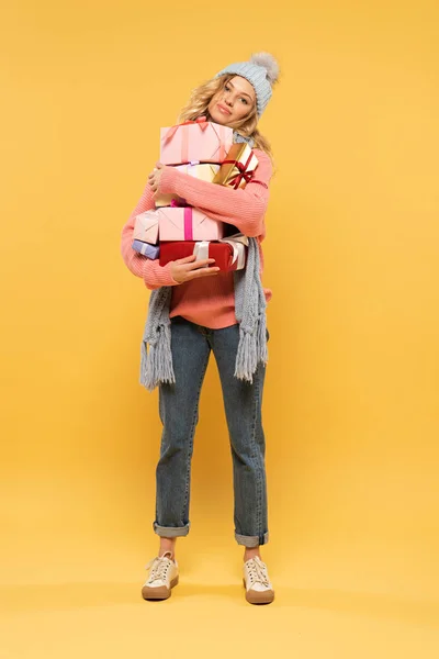 Jovem mulher de chapéu e cachecol segurando caixas de presente no fundo amarelo — Fotografia de Stock