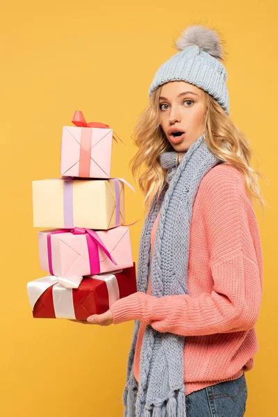 Shocked woman in hat and scarf holding gift boxes isolated on yellow — Stock Photo