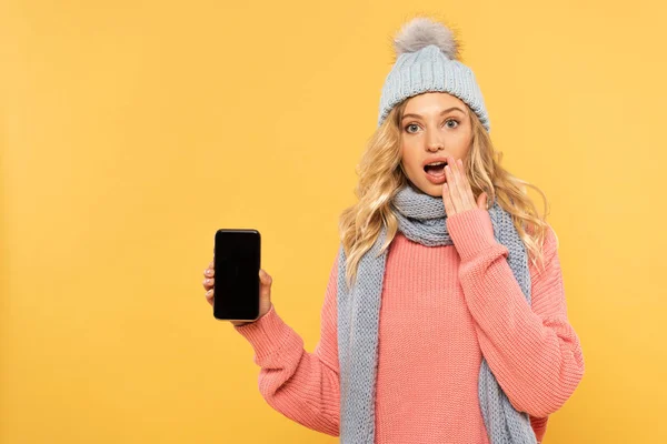 Femme surprise en chapeau et écharpe tenant smartphone avec écran blanc isolé sur jaune — Photo de stock