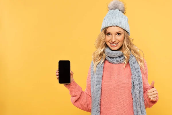 Smiling woman in hat holding smartphone with blank and showing approval sign isolated on yellow — Stock Photo