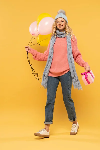 Mujer sonriente en sombrero y bufanda sosteniendo globos y caja de regalo sobre fondo amarillo - foto de stock