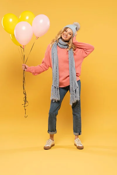 Lächelnde Frau mit Hut und Schal mit Luftballons auf gelbem Hintergrund — Stockfoto