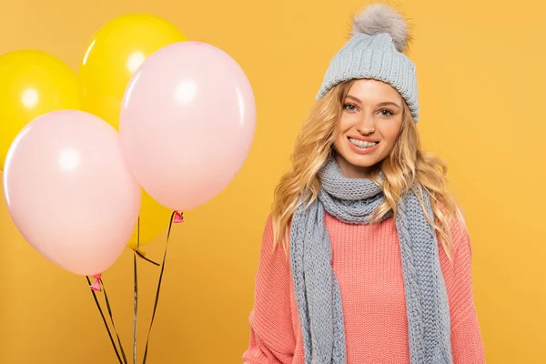 Donna in cappello e sciarpa sorridente e con palloncini isolati su giallo — Foto stock