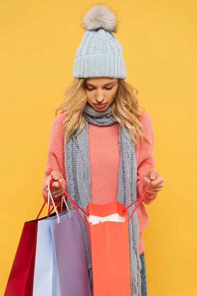 Woman in hat and scarf looking in shopping bags isolated on yellow — Stock Photo