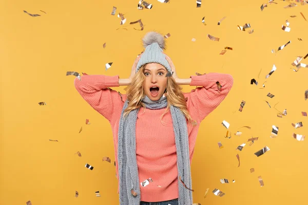 Surprised woman in hat and scarf with falling golden confetti isolated on yellow — Stock Photo