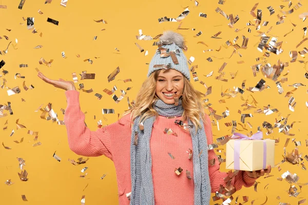 Mujer sonriente en sombrero sosteniendo caja de regalo mientras cae confeti dorado aislado en amarillo - foto de stock