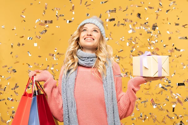 Femme heureuse dans le chapeau tenant boîte cadeau et sacs à provisions sous confettis tombant isolé sur jaune — Photo de stock