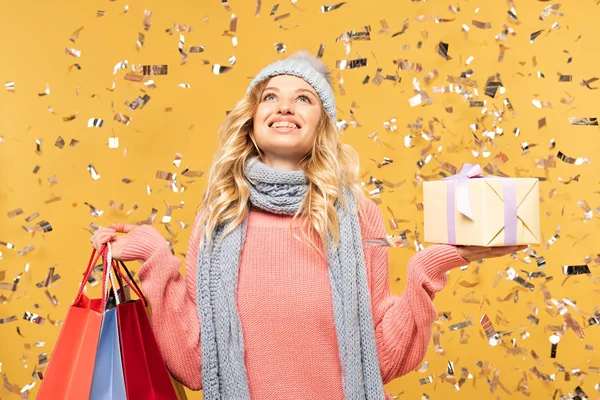 Happy woman in hat holding gift box and shopping bags under falling confetti isolated on yellow — Stock Photo