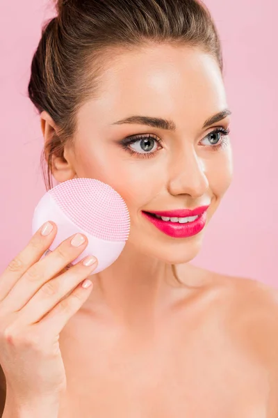 Smiling naked beautiful woman with pink lips holding facial cleansing brush isolated on pink — Stock Photo