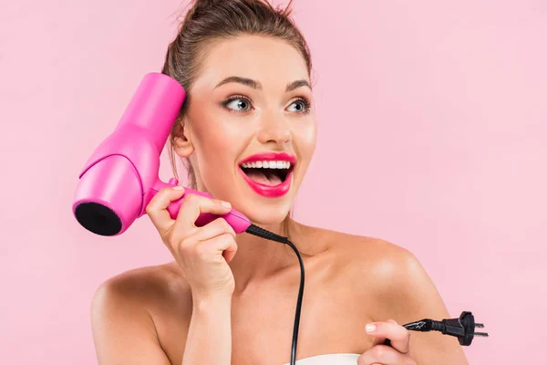 Aufgeregt schöne Frau mit offenem Mund und rosa Lippen hält Haartrockner isoliert auf rosa — Stockfoto