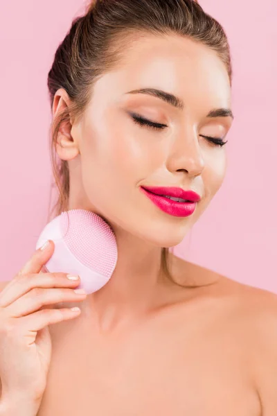 Довольная обнаженная красивая женщина с розовыми губами с помощью щетки для очищения лица с закрытыми глазами, изолированными на розовый — стоковое фото
