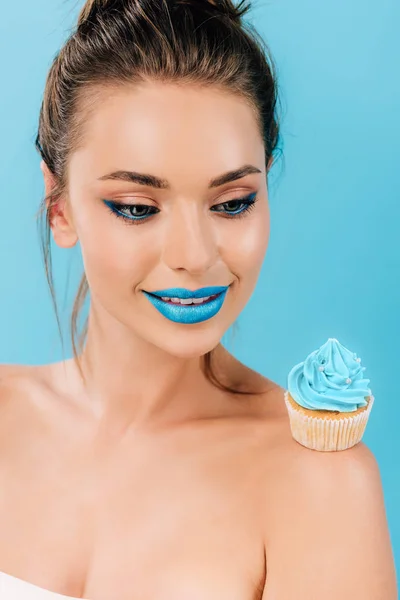 Smiling naked beautiful woman with blue lips looking at cupcake on shoulder isolated on blue — Stock Photo