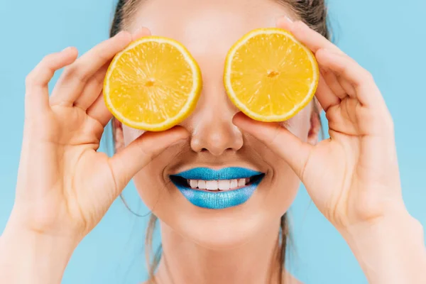 Smiling beautiful woman with blue lips holding orange halves on eyes isolated on blue — Stock Photo
