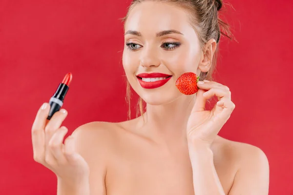 Hermosa mujer desnuda con labios rojos sosteniendo lápiz labial y fresa aislado en rojo - foto de stock