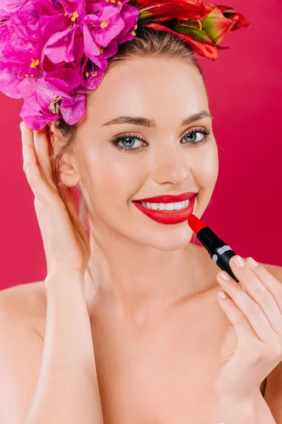 Hermosa mujer desnuda sonriente con labios rojos y corona floral en la cabeza aplicando lápiz labial aislado en carmesí - foto de stock