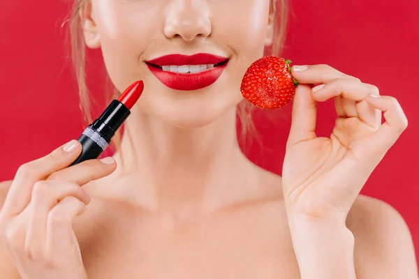 Cropped view of naked beautiful woman with red lips holding lipstick and strawberry isolated on red — Stock Photo