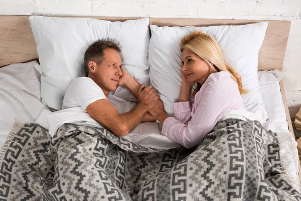 Vue du dessus du couple souriant tenant la main tout en étant couché au lit — Photo de stock