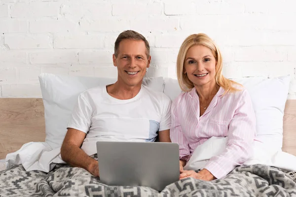 Усміхнена зріла пара з ноутбуком дивиться в камеру в ліжку — стокове фото