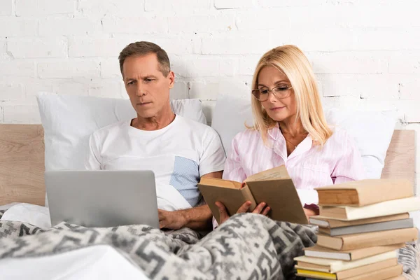 Hombre usando el ordenador portátil mientras su esposa lee el libro en la cama - foto de stock