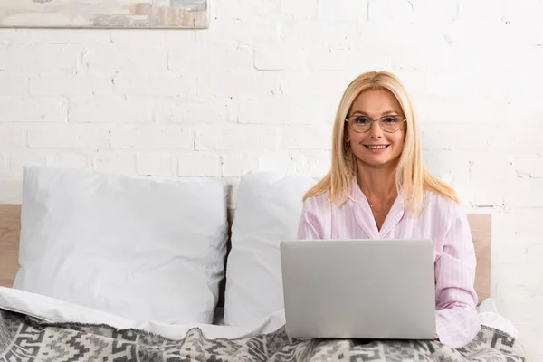 Mulher sorridente em óculos olhando para a câmera enquanto usa laptop na cama — Fotografia de Stock