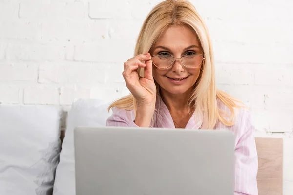 Mujer sonriente con anteojos usando laptop en la cama - foto de stock