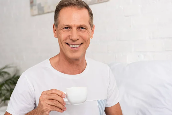Мужчина держит чашку кофе и улыбается перед камерой в спальне — стоковое фото