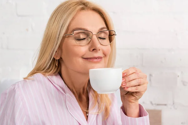Femme souriante dans les lunettes sentant le café dans la tasse à la maison — Photo de stock