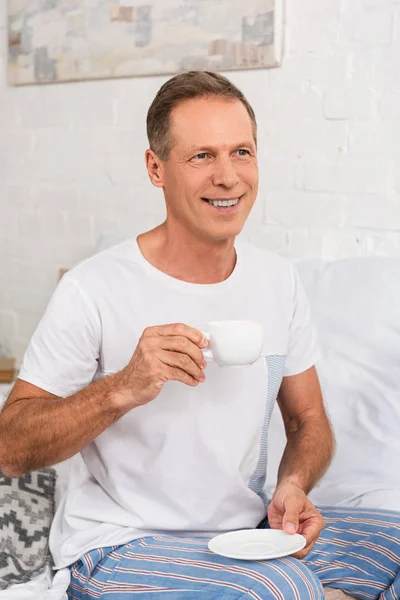 Uomo sorridente bere caffè mentre seduto sul letto — Foto stock