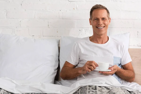 Чоловік тримає чашку кави в ліжку і посміхається на камеру — стокове фото