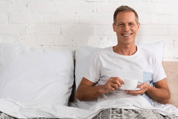 Uomo che tiene una tazza di caffè a letto e sorride alla macchina fotografica — Foto stock