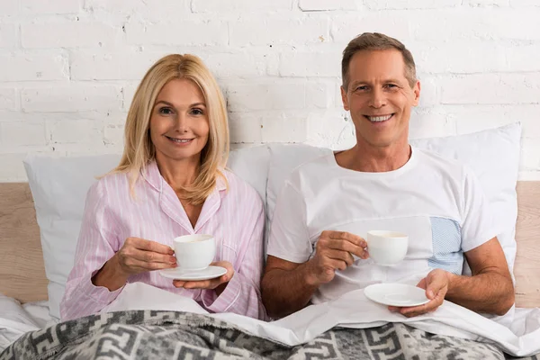 Зрелая пара с кофейными чашками, улыбающаяся в постели перед камерой — стоковое фото