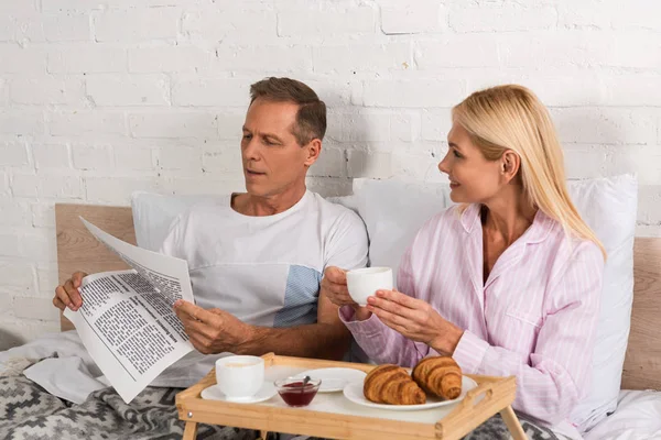 Hombre leyendo el periódico mientras desayuna con su esposa en la cama - foto de stock