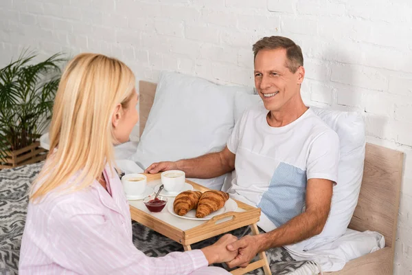 Couple souriant tenant la main tout en prenant le petit déjeuner au lit — Photo de stock