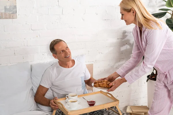 Woman giving croissants to husband with breakfast tray in bed — Stock Photo