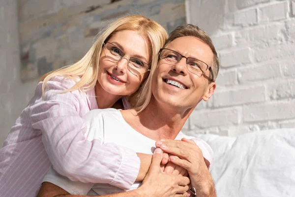 Улыбающаяся пара в пижаме и очках обнимается в спальне по утрам — стоковое фото