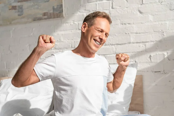 Homem sorridente que se estende enquanto está sentado na cama de manhã — Fotografia de Stock