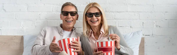 Laughing couple in 3d glasses holding buckets with popcorn on bed, panoramic shot — Stock Photo