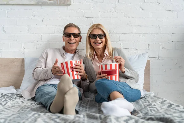 Smiling couple in 3d glasses with popcorn buckets on bed — Stock Photo