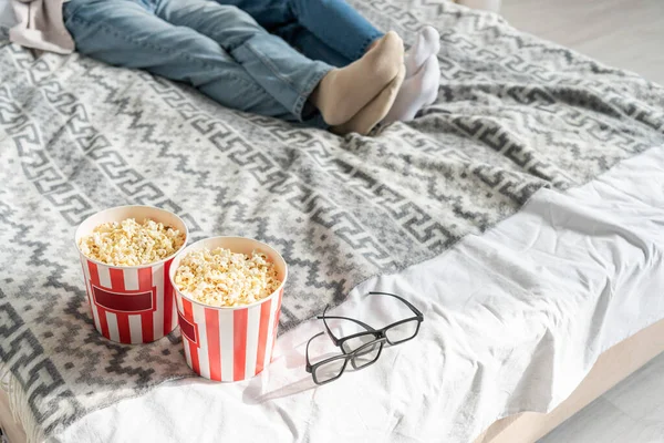 Vista ritagliata di coppia sdraiata sul letto accanto a secchi di popcorn e bicchieri 3d — Foto stock