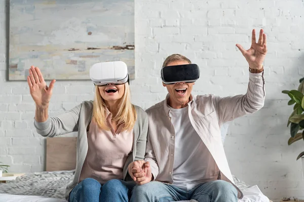 Aufgeregtes Paar in Virtual-Reality-Headsets, die Hände im Bett haltend — Stockfoto