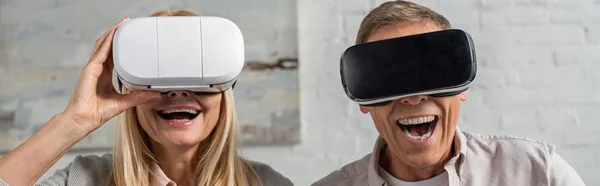 Панорамный снимок смеющейся пары в наушниках виртуальной реальности дома — стоковое фото