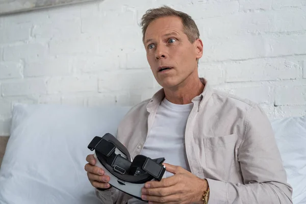 Hombre sorprendido sosteniendo auriculares de realidad virtual en la cama - foto de stock