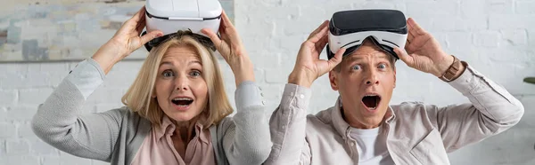 Panoramic shot of shocked couple with virtual reality headsets looking at camera — Stock Photo