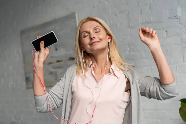 Smiling woman with smartphone and earphones dancing at home — Stock Photo