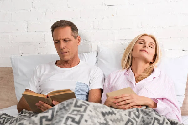 Man reading book beside wife sleeping in bed — Stock Photo